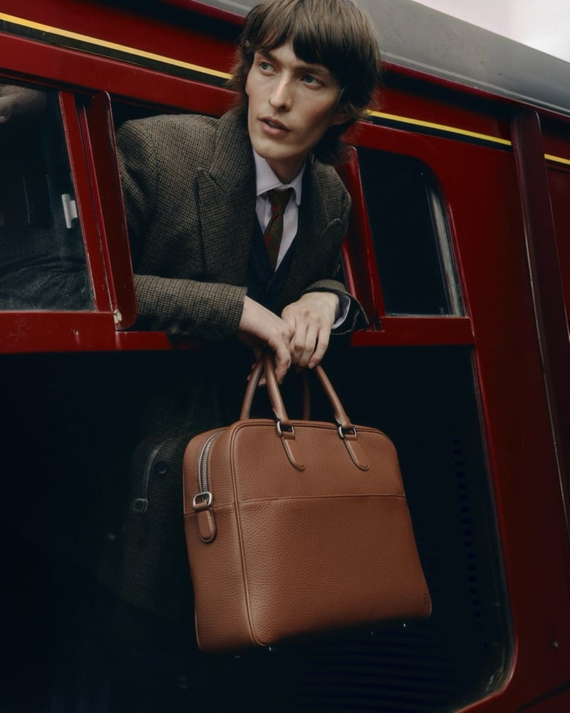 Erik Kettschick leans out of a train window in a checked tweed jacket and striped tie, holding a cognac leather briefcase, for Carl Friedrik's fall 2026 campaign