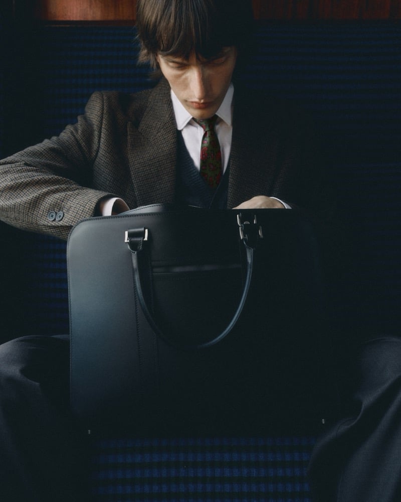 Erik Kettschick sits in a train compartment in a checked jacket and floral tie, holding a leather briefcase, for Carl Friedrik's fall 2026 campaign