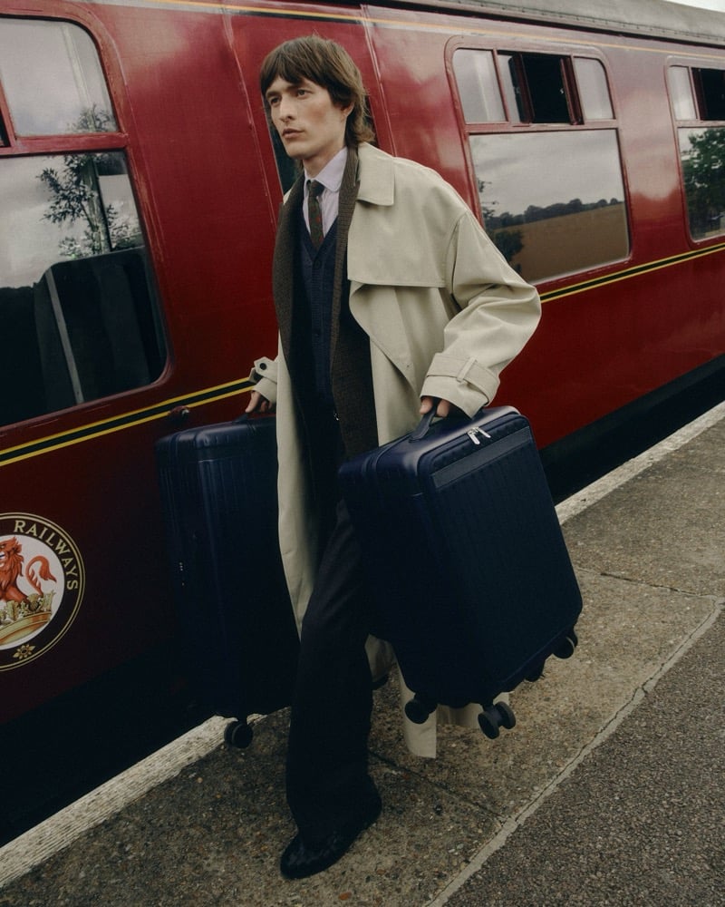 Erik Kettschick walks along a railway platform in a beige trench coat and scarf, carrying two hard-shell suitcases, beside a vintage red railway carriage for Carl Friedrik's fall 2026 campaign