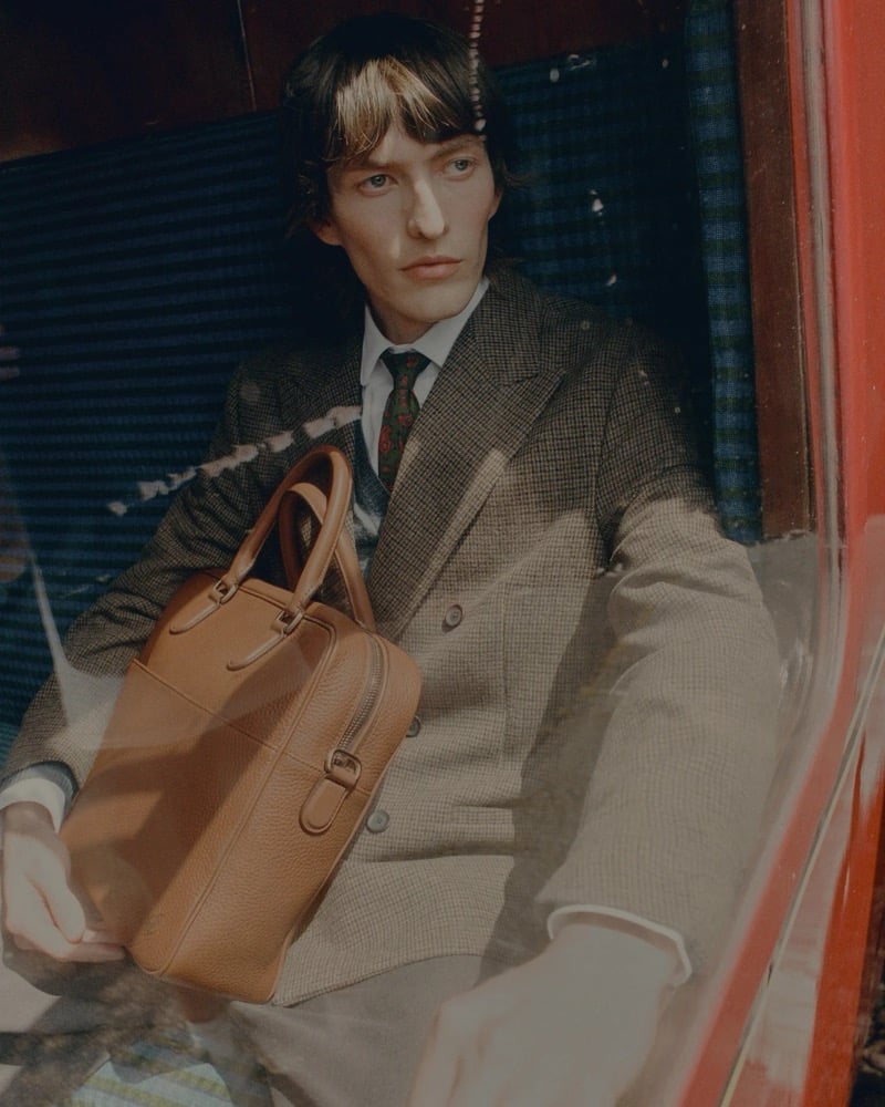 Erik Kettschik seen through a train window in a checked jacket, holding a cognac leather briefcase, for Carl Friedrik's fall 2026 campaign