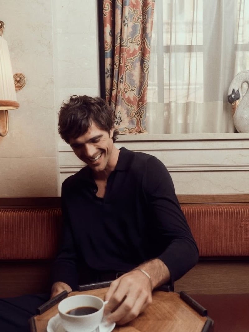 Jacob laughs over a cup of coffee in a black top, the Cartier LOVE Unlimited bracelet visible on his wrist.