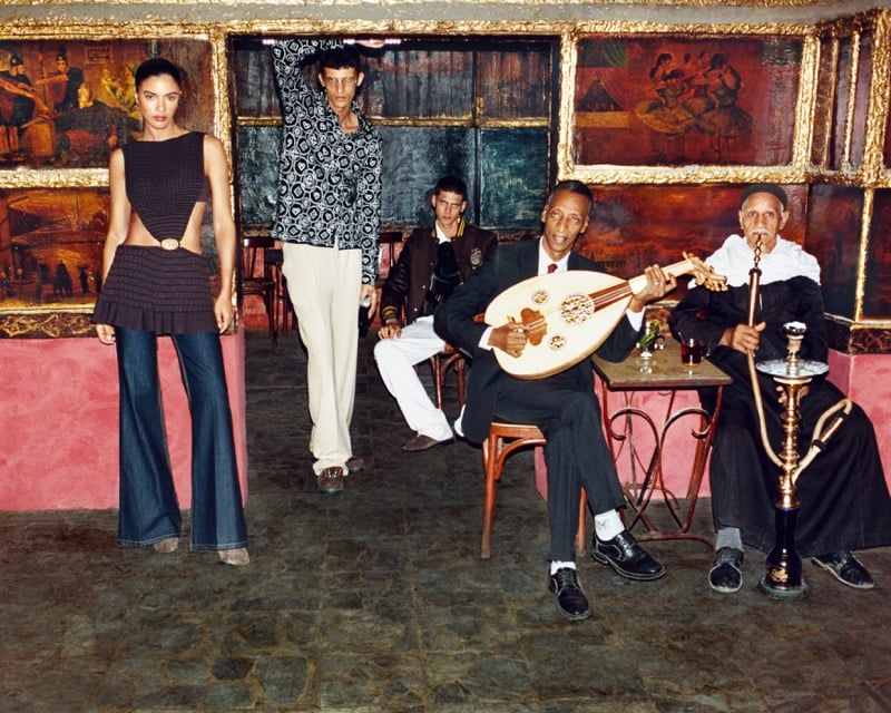 Rania Benchegra, Youssef Rocha, and Youssef Sharaf standing and seated inside a traditional Cairo cafe, with two local men playing an oud and smoking a hookah in the background