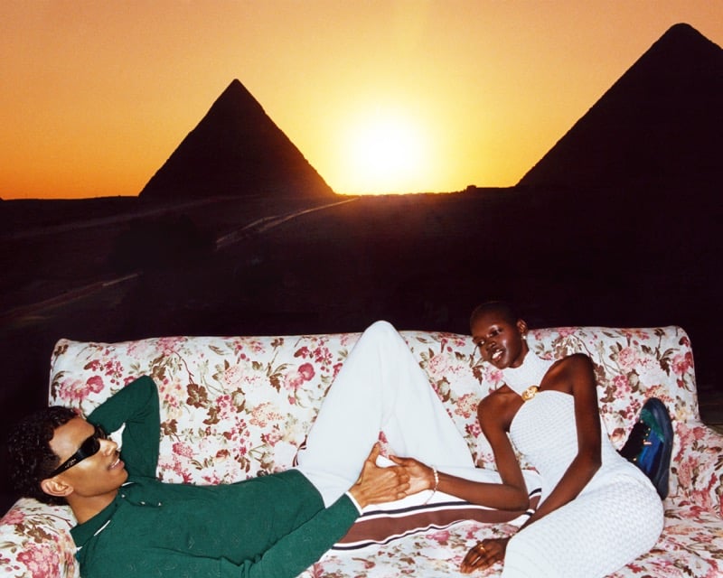 Mohamed Hassan lying on a floral sofa in sunglasses and a green sweater as Adhar Abiem sits beside him, the Pyramids of Giza silhouetted at sunset behind them
