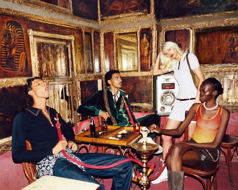 Lulu Wood stands over three other campaign models gathered around a game table inside an ornate Cairo lounge decorated with pharaonic murals