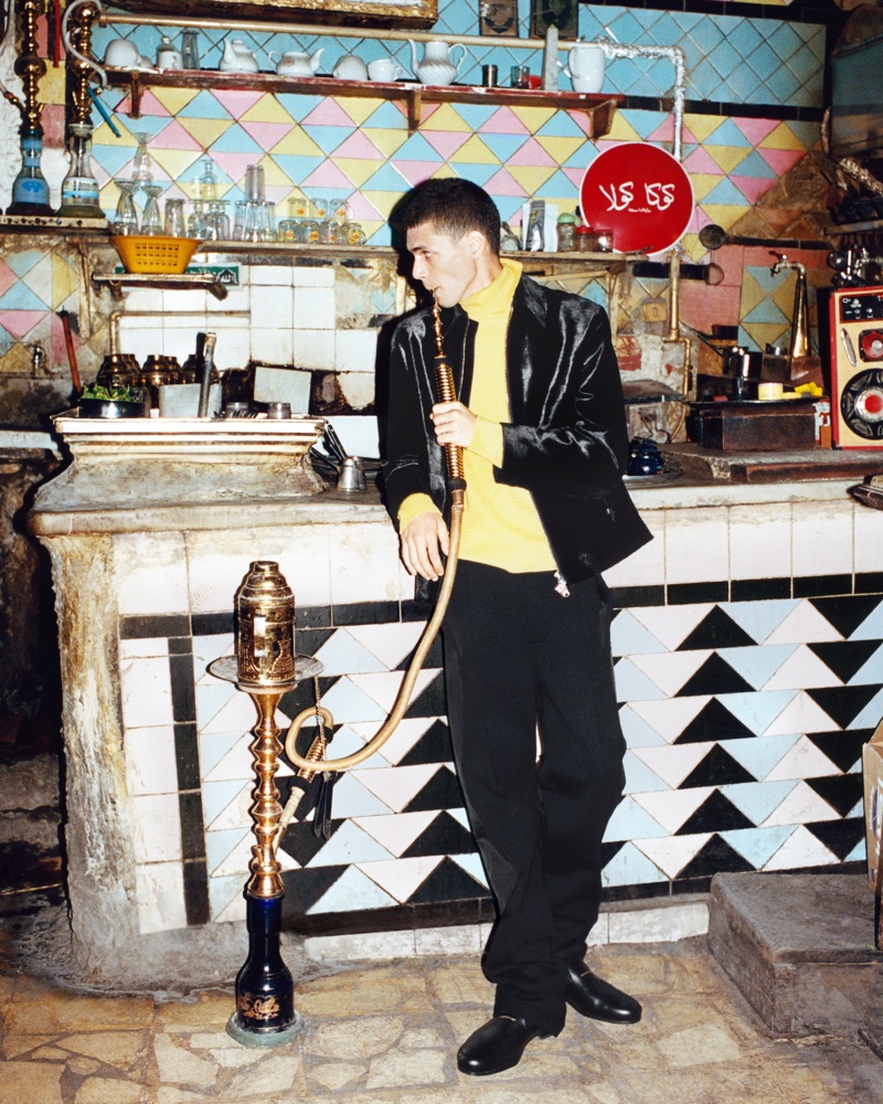 Youssef Sharaf in a yellow turtleneck and black velvet jacket lighting a hookah at a tiled Cairo cafe counter