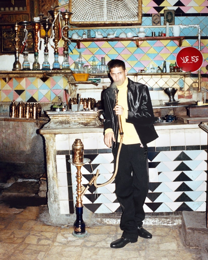 Youssef Sharaf in a yellow turtleneck and black jacket, holding a hookah pipe in a cafe with mosaic-tiled walls