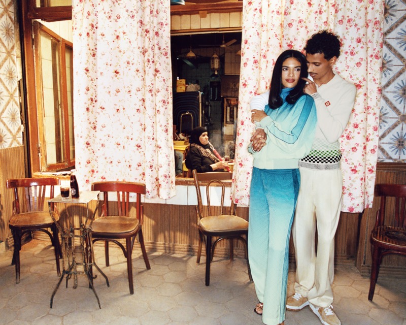 Two Casablanca campaign models embrace in a cafe doorway framed by floral curtains, a local woman seated inside