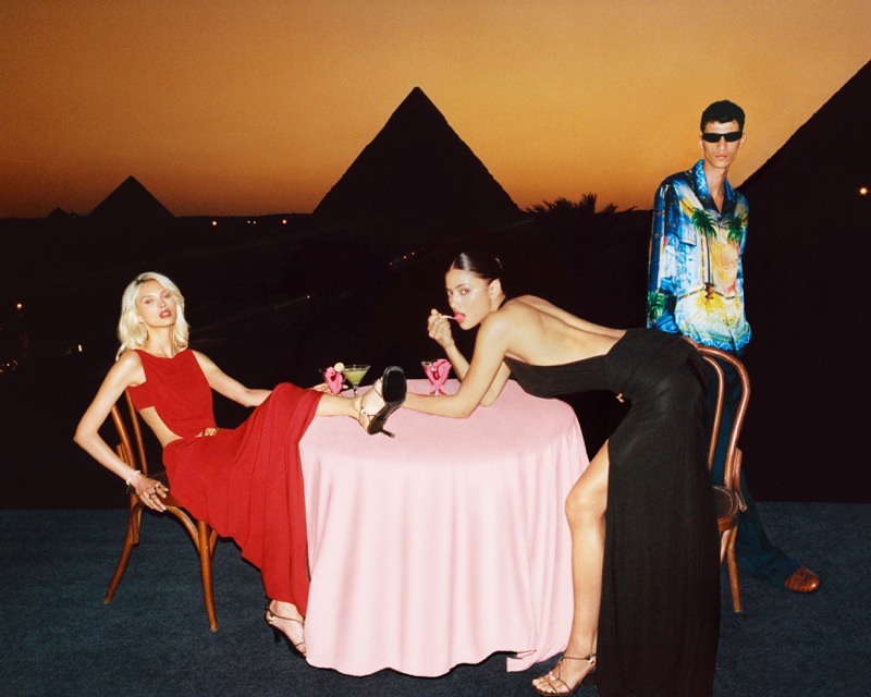 Lulu Wood and Rania Benchegra sit at a table with cocktails as a third model stands behind them, the Pyramids of Giza silhouetted at sunset