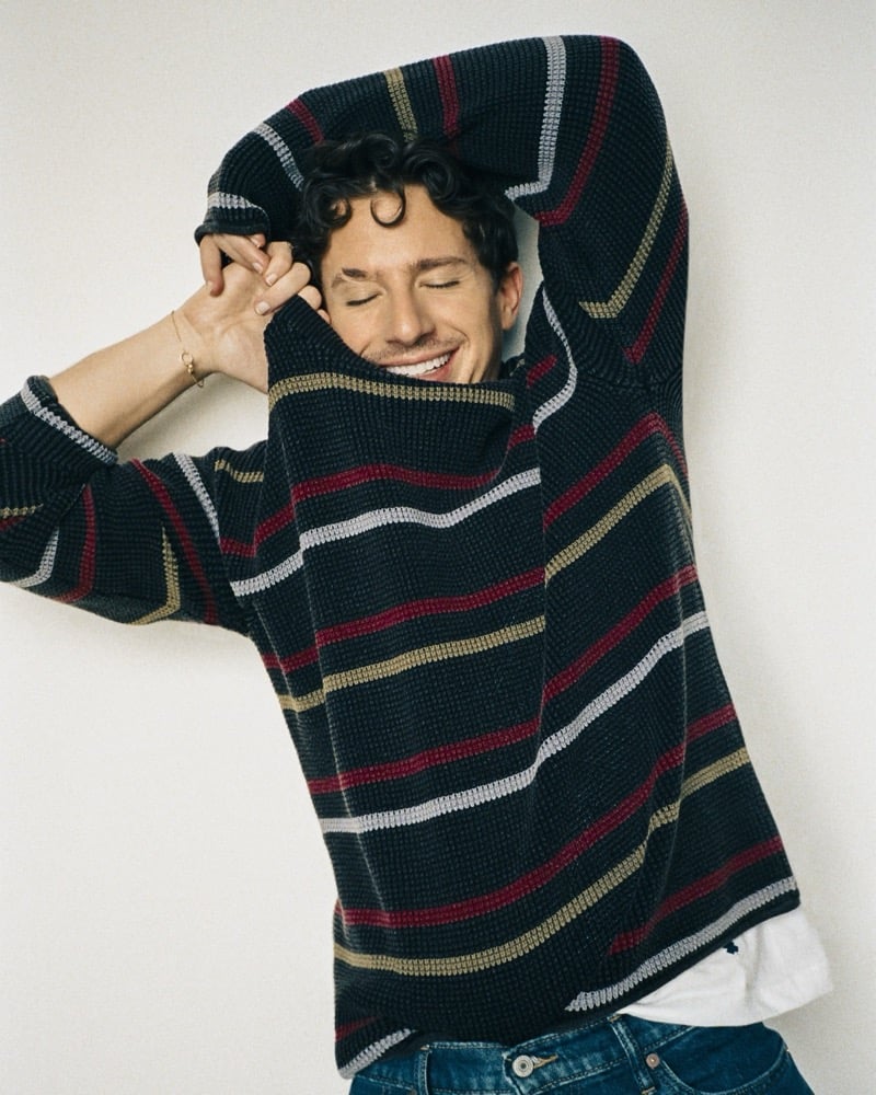 Charlie Puth laughs with his eyes closed while pulling a striped knit sweater over his face, arm raised overhead