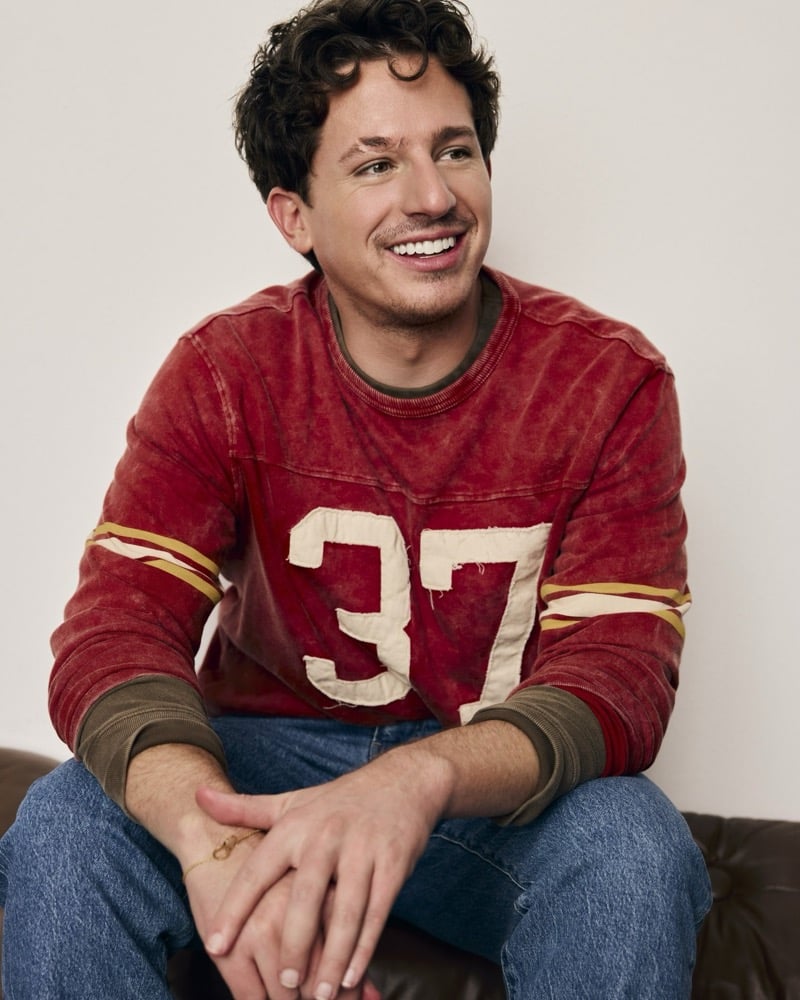 Charlie Puth smiles while seated in a faded red football jersey numbered 37 over blue jeans