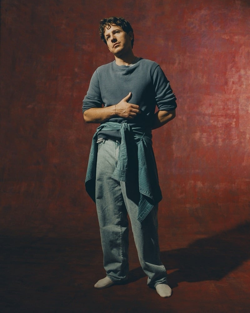 Charlie Puth stands in a gray thermal top with a striped shirt tied at his waist over wide jeans, against a red backdrop