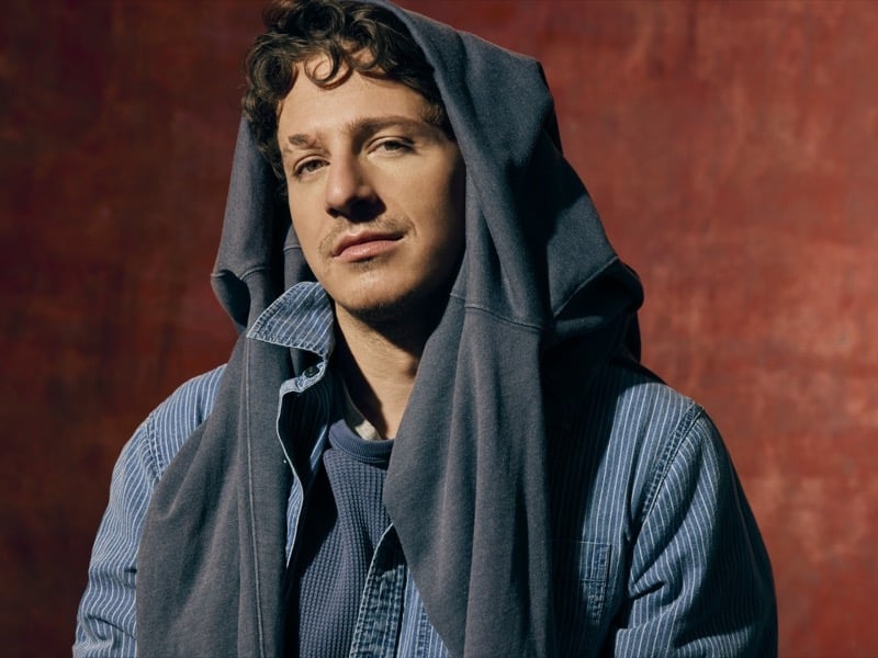 Charlie Puth pulls a gray hoodie over his head, a striped denim shirt visible underneath, against a red backdrop