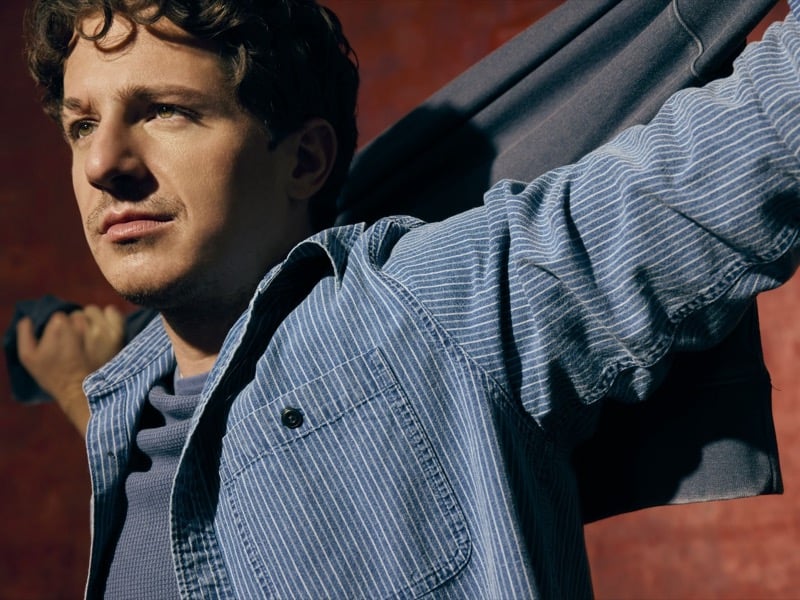 Close-up of Charlie Puth in a striped denim shirt jacket, arm raised, against a red backdrop