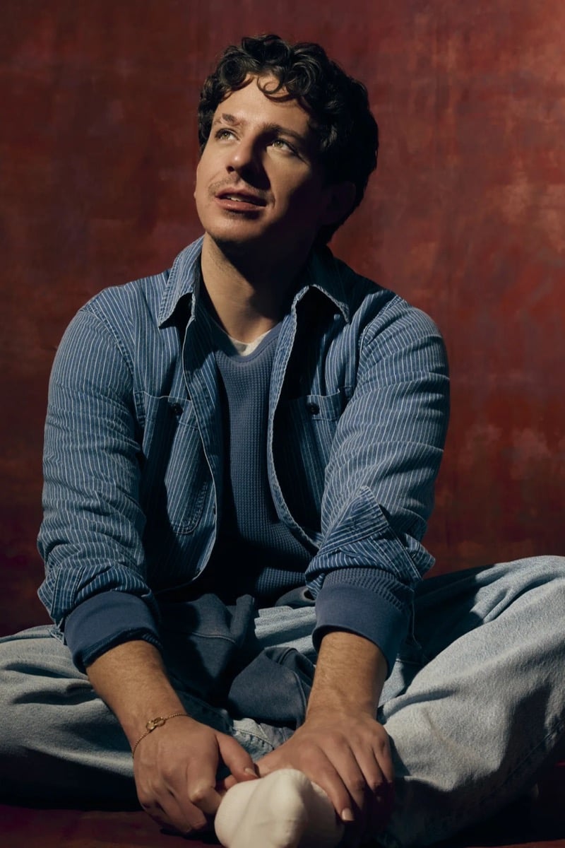 Charlie Puth sits cross-legged in a striped denim shirt jacket over a thermal top and jeans against a red backdrop