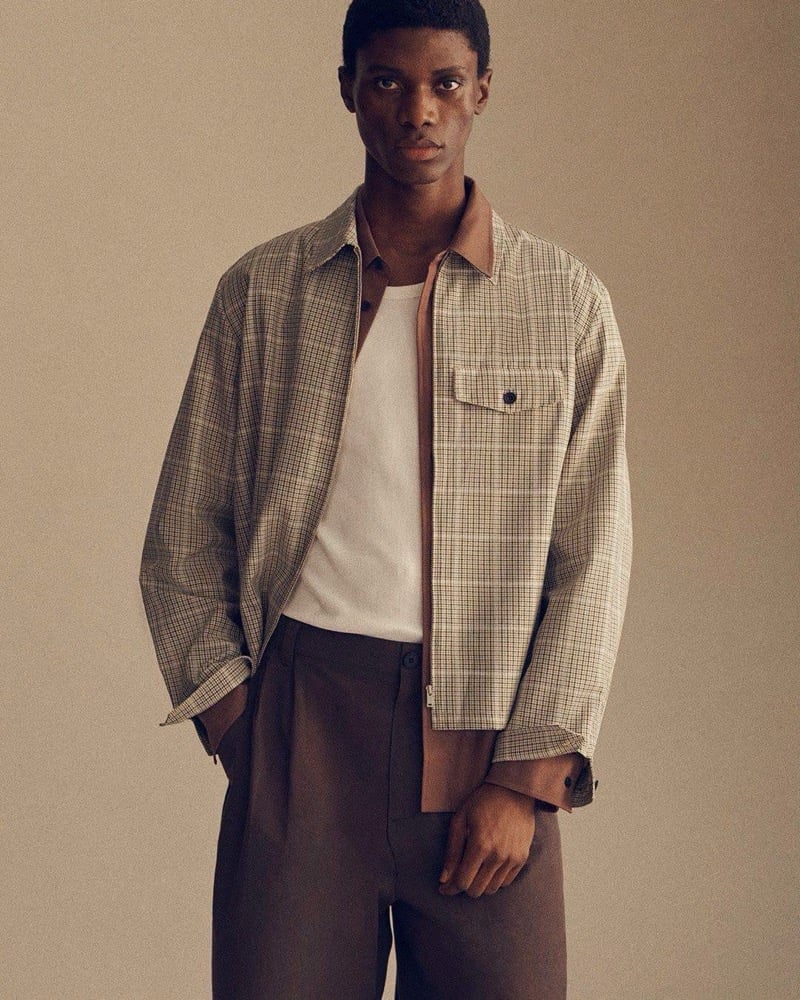 Awwal Adeoti wears an open plaid zip jacket with tan trim over a tee and pleated brown trousers