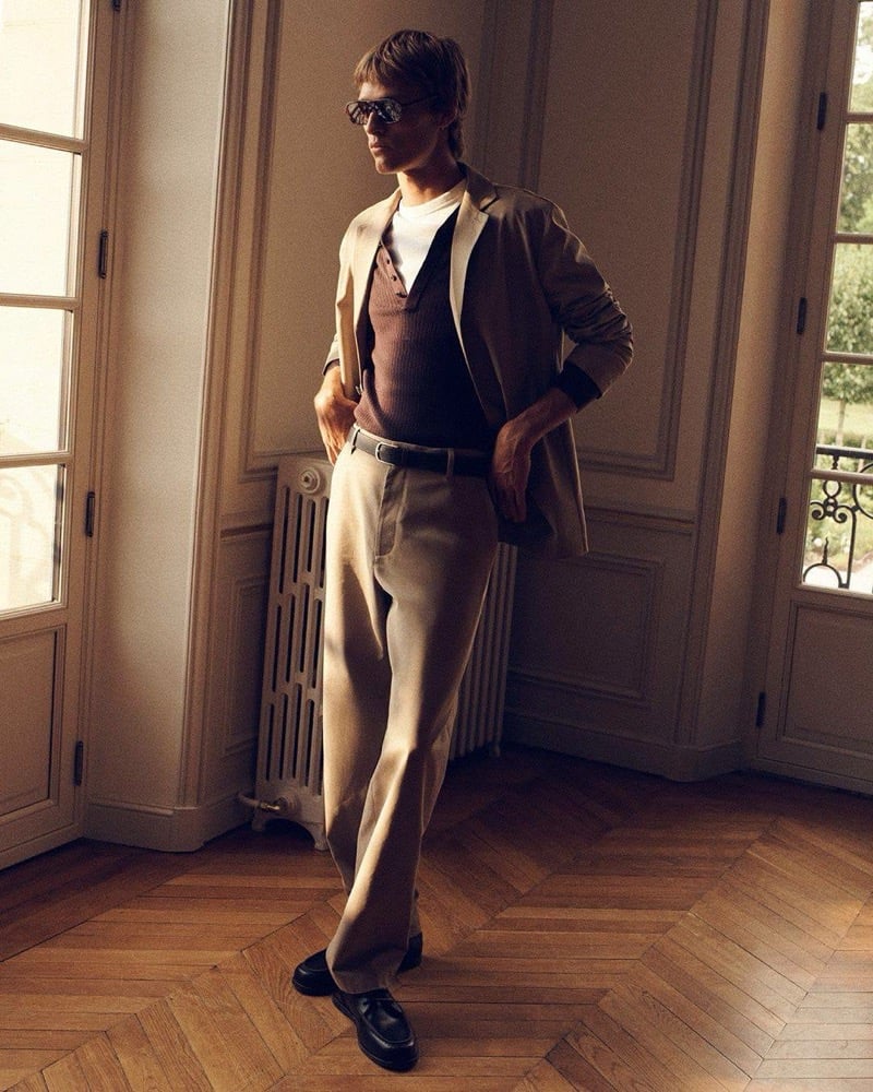 Yeray Allgayer wears a tan blazer over a brown knit henley with beige trousers and sunglasses, standing before French doors