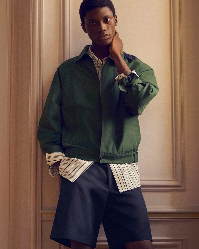 Awwal Adeoti wears an olive green zip jacket over a striped shirt with dark tailored shorts