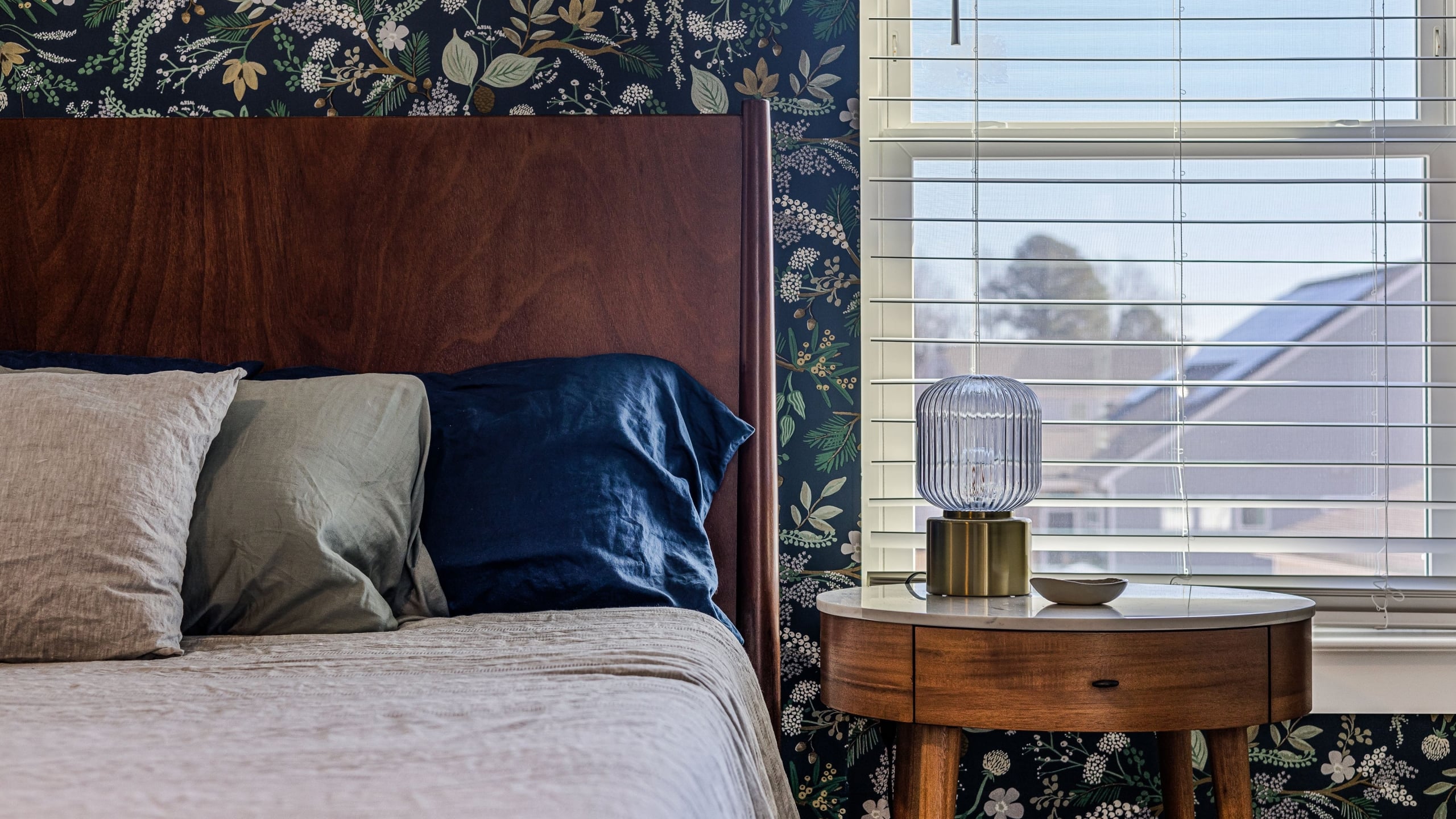 A walnut bed headboard sits against a dark botanical wallpaper beside a window