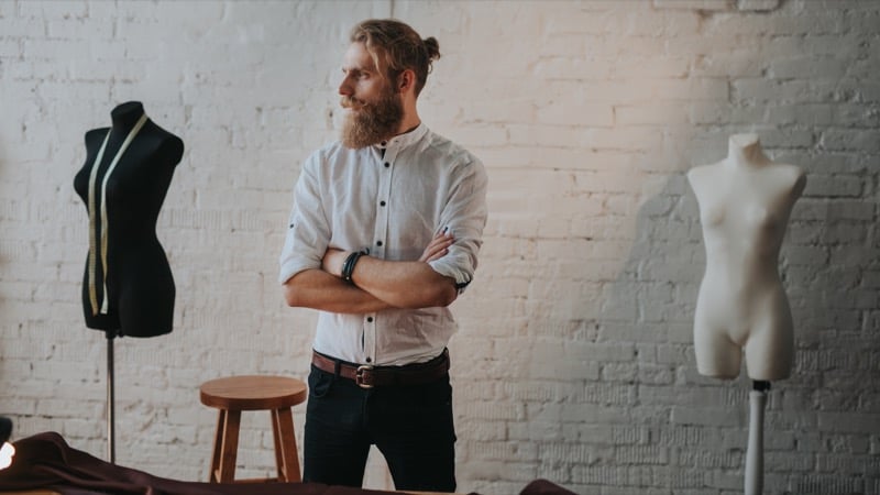 A bearded man with his hair in a bun stands with arms crossed in a tailoring studio, dress forms and a tape measure visible around him