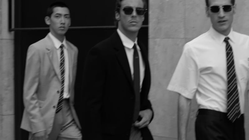 Three men in Dior tailoring walk in black-and-white motion blur, trousers and shoes visible beneath a light suit, a black suit and a white shirt