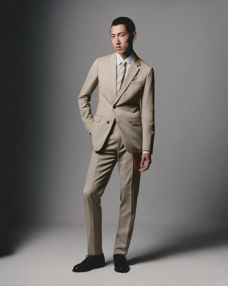 A model in a pale suit and striped tie stands with one hand in his pocket against a gray studio backdrop