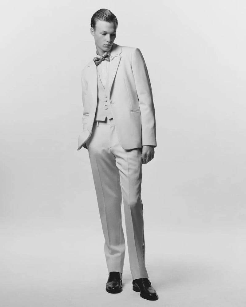 A model looks down in a light-colored tuxedo with a vest and patterned bow tie against a white backdrop