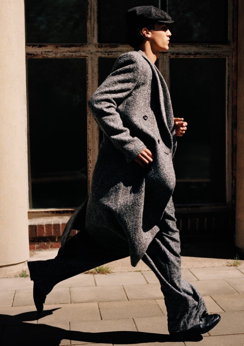 Morgan Sahadow walking mid-stride in a long herringbone coat, wide-leg trousers, and a flat cap for Emporio Armani's fall-winter 2026 campaign