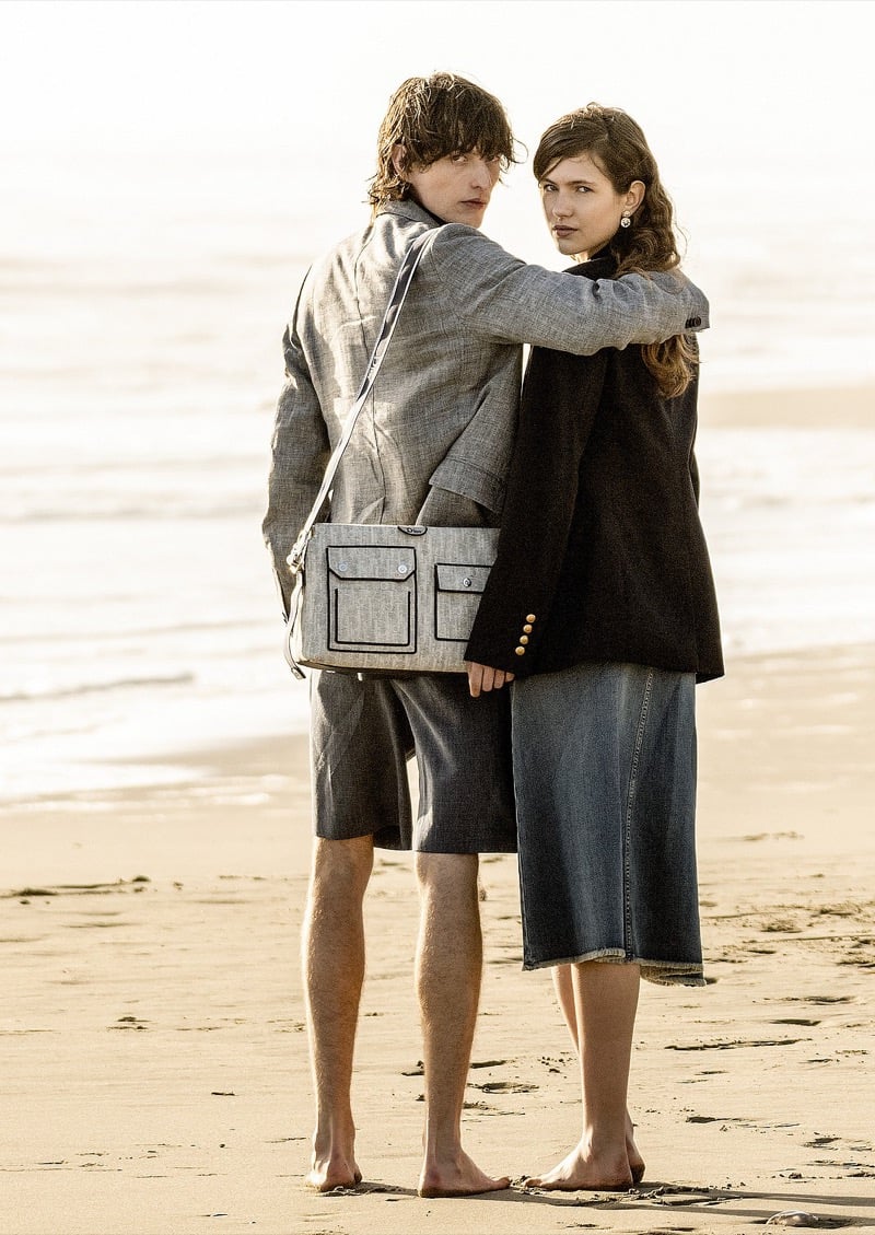 Erik Kettschick stands on the beach with a female model at his shoulder, wearing a gray blazer with a matching Dior bag across his back