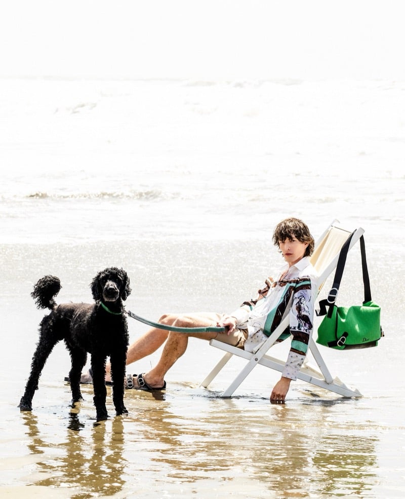 Erik Kettschick reclines in a beach chair in a printed shirt and shorts, holding a poodle's leash beside a green Dior bag
