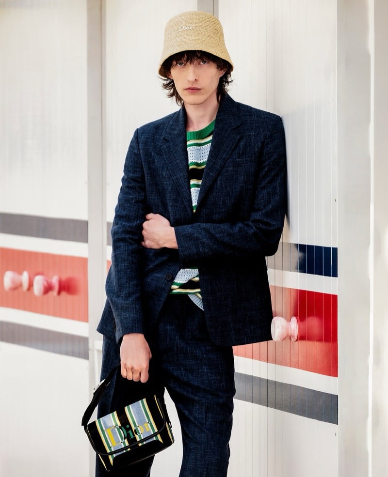 Erik Kettschick leans against a striped bathing cabin in a heathered suit, striped sweater, and straw Dior bucket hat