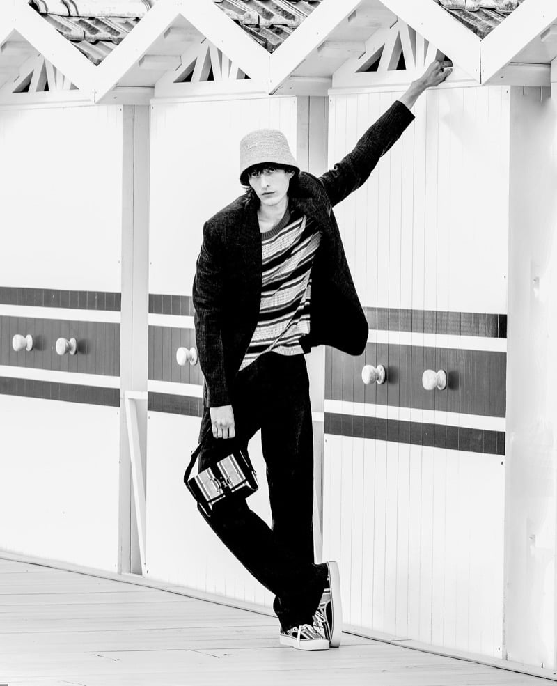Erik Kettschick reaches upward against a bathing cabin in a black-and-white portrait, wearing a textured suit, striped sweater, and bucket hat