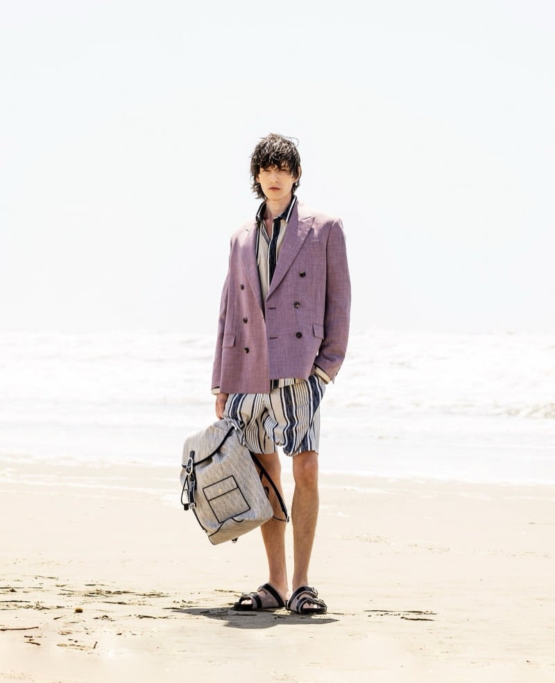 Erik Kettschick stands barefoot on the shoreline in a mauve double-breasted blazer and striped shorts, holding a canvas Dior bag