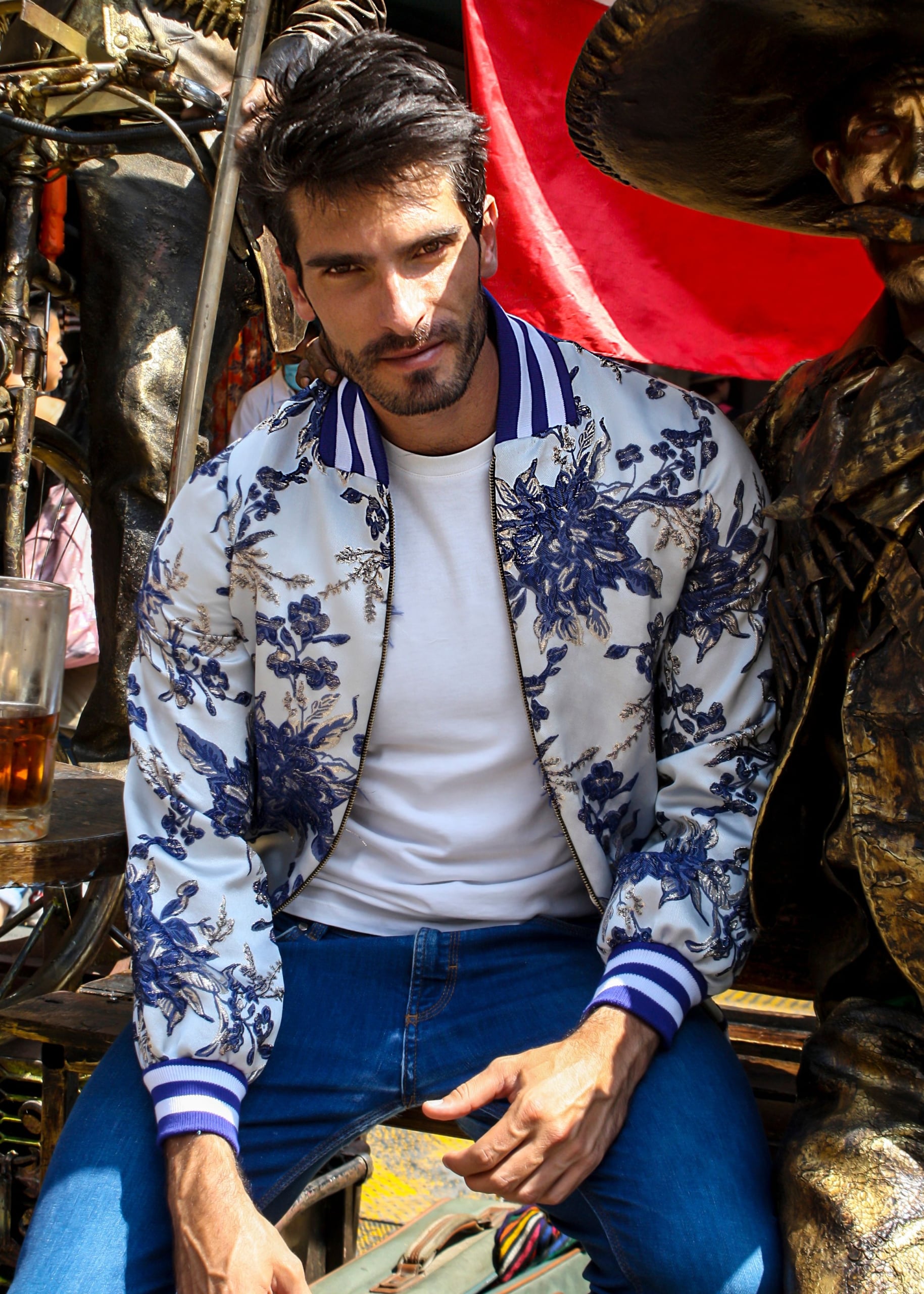 A man sits outdoors wearing a navy floral bomber jacket over a white t-shirt