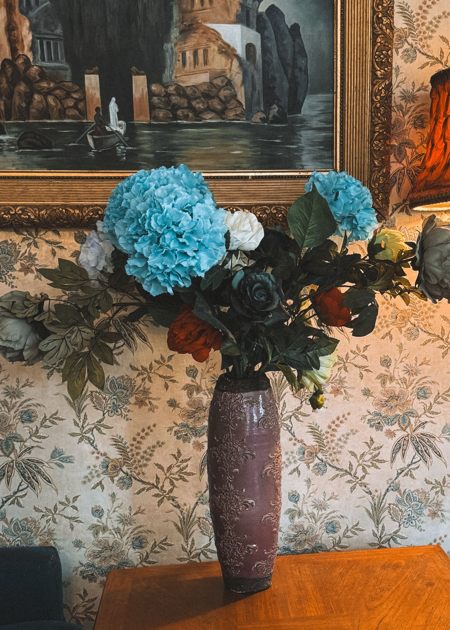 A vase of flowers sits on a table in front of a wall covered in pale floral wallpaper and framed art