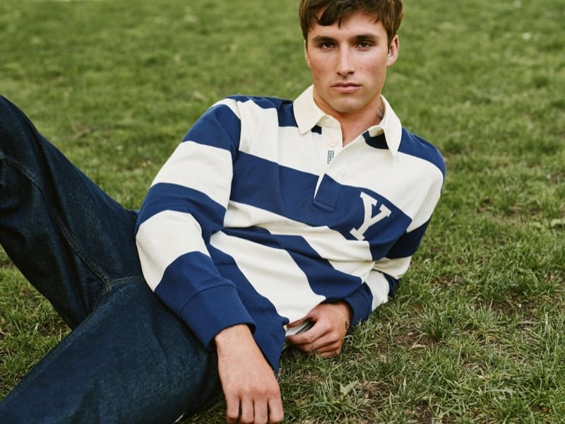 Jack Lumsden reclining on the grass in a striped rugby shirt with a Y patch and wide-leg jeans