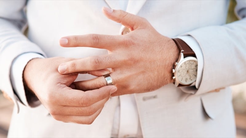 A groom in a light suit adjusts his silver wedding band outdoors, watch visible on his wrist