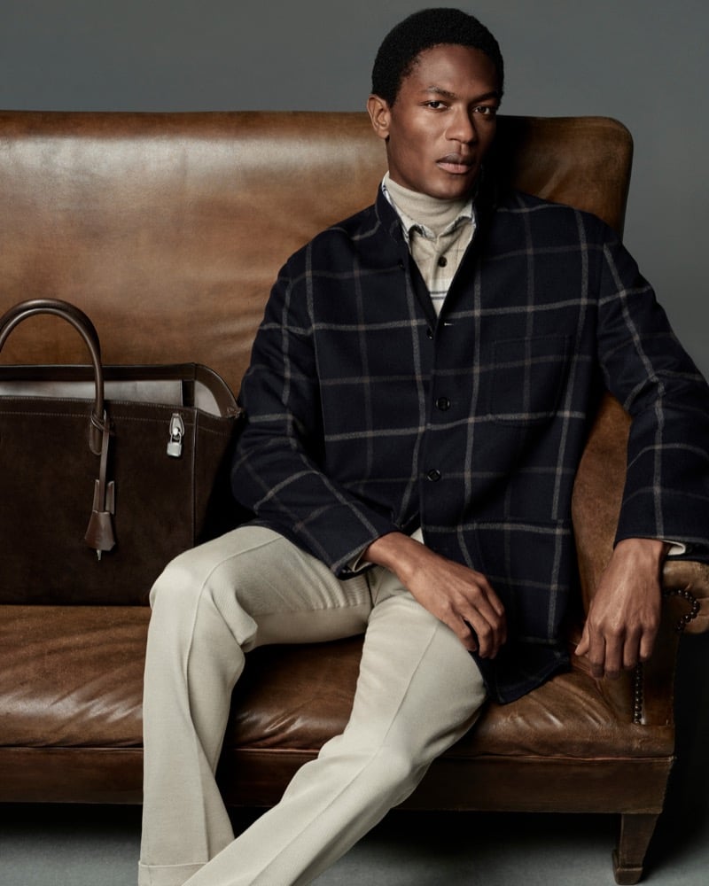 Hamid Onifade wears a dark windowpane check jacket with cream trousers for Dunhill's fall-winter 2026 campaign, a leather tote beside him