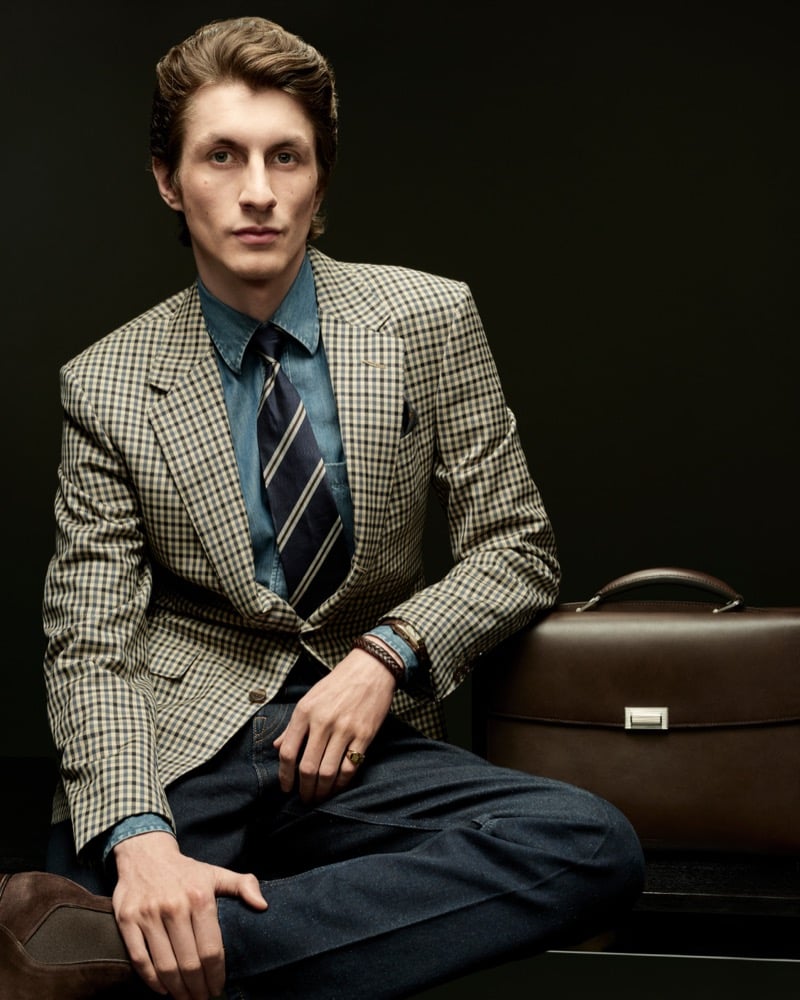 Henry Kitcher wears a checked blazer with a denim shirt, striped tie and jeans beside a brown leather briefcase for Dunhill's fall-winter 2026 campaign