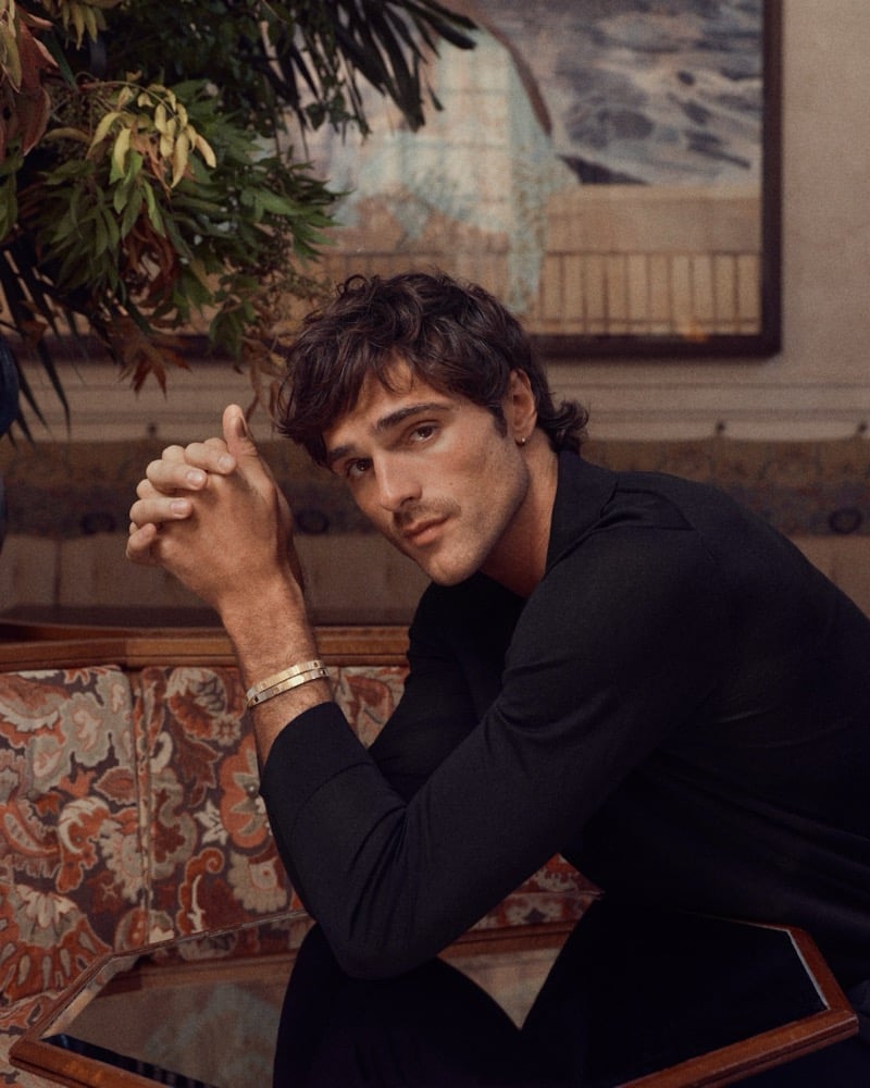 Jacob wears a black top with the Cartier LOVE Unlimited bracelet, hands clasped together in a decorated interior.