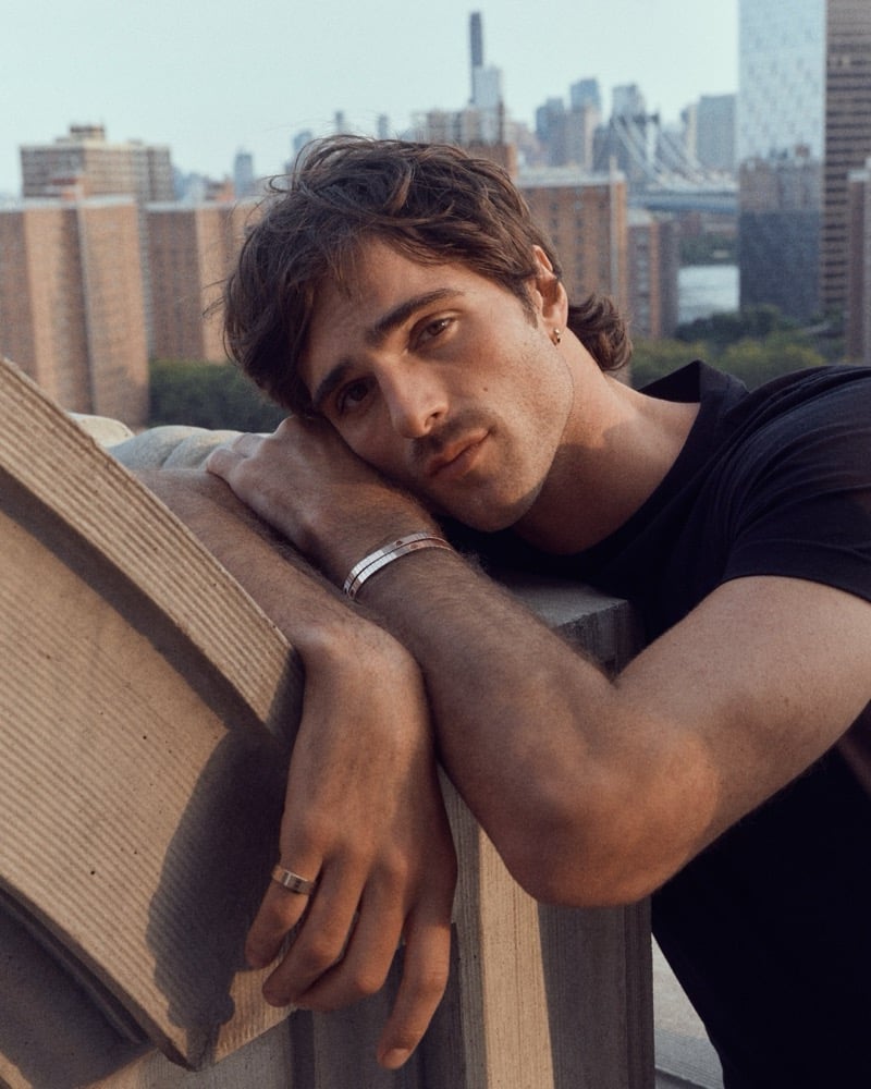 Jacob rests his head on his crossed arms in a black T-shirt with the Cartier LOVE Unlimited bracelet, the New York skyline behind him.