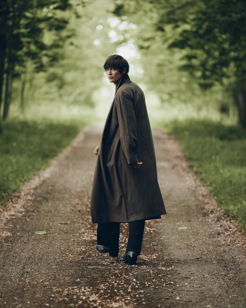 Alfie Nickerson in a brown trench coat looking back over his shoulder while walking down a tree-lined path