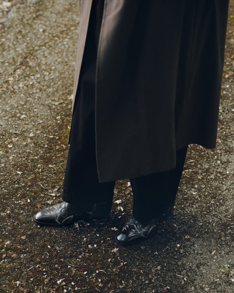Close-up of black leather Wilder boots with floral laser-cut appliqué beneath the hem of a brown trench coat