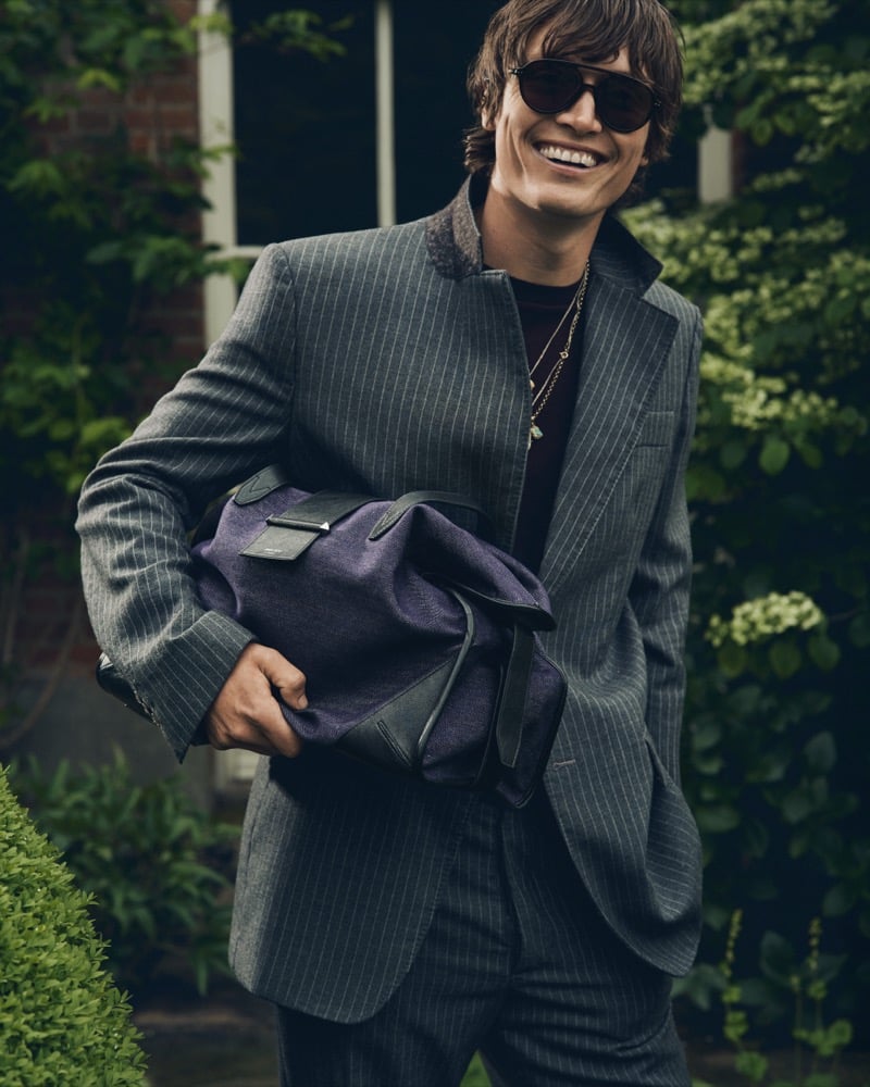 Alfie Nickerson laughing in a pinstripe suit and sunglasses while holding a deep purple Jimmy Choo Bar holdall