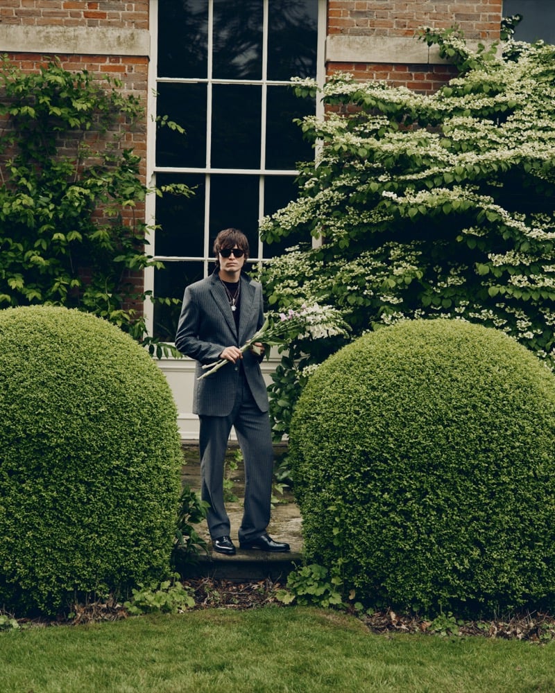 Alfie Nickerson in a pinstripe suit holding a bouquet of pink and white flowers between two round topiary hedges