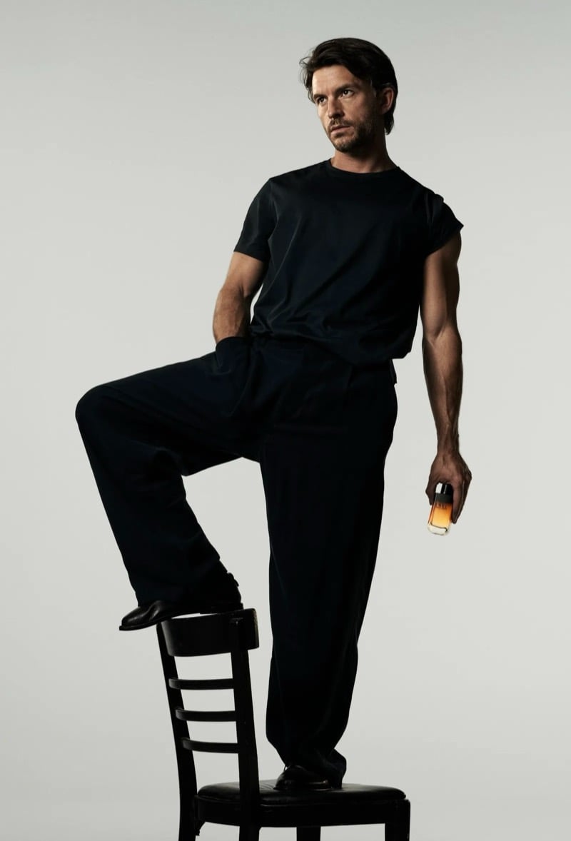Jonathan Bailey in a black T-shirt and relaxed black trousers standing with one foot propped on a chair, holding the Giorgio Armani I Will bottle