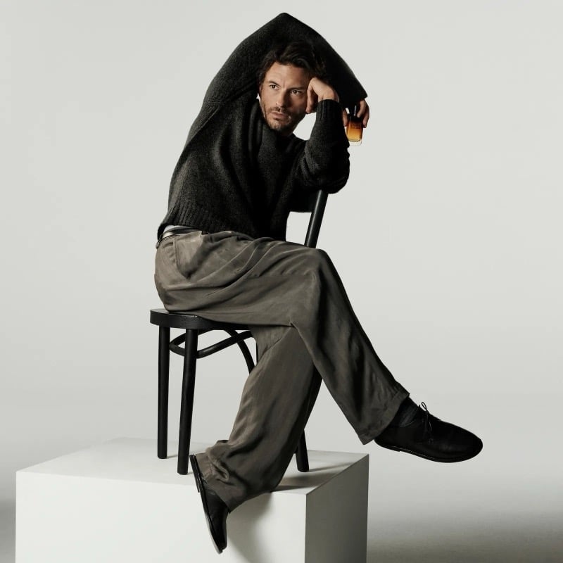 Jonathan Bailey in a dark knit sweater and olive-toned trousers seated on a chair atop a white pedestal, holding the Giorgio Armani I Will bottle