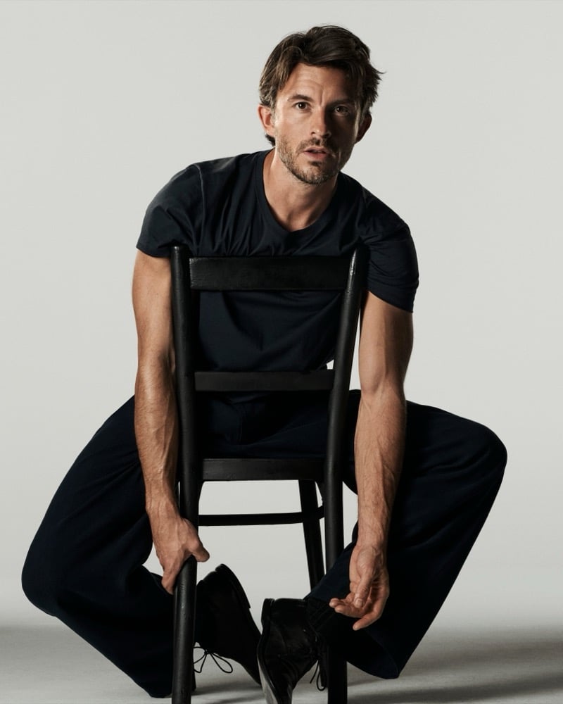 Jonathan Bailey in a black T-shirt sitting backward on a chair with his arms resting on the chair back