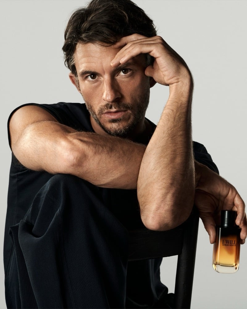 Jonathan Bailey in a sleeveless black top leaning on a chair back, holding the Giorgio Armani I Will bottle