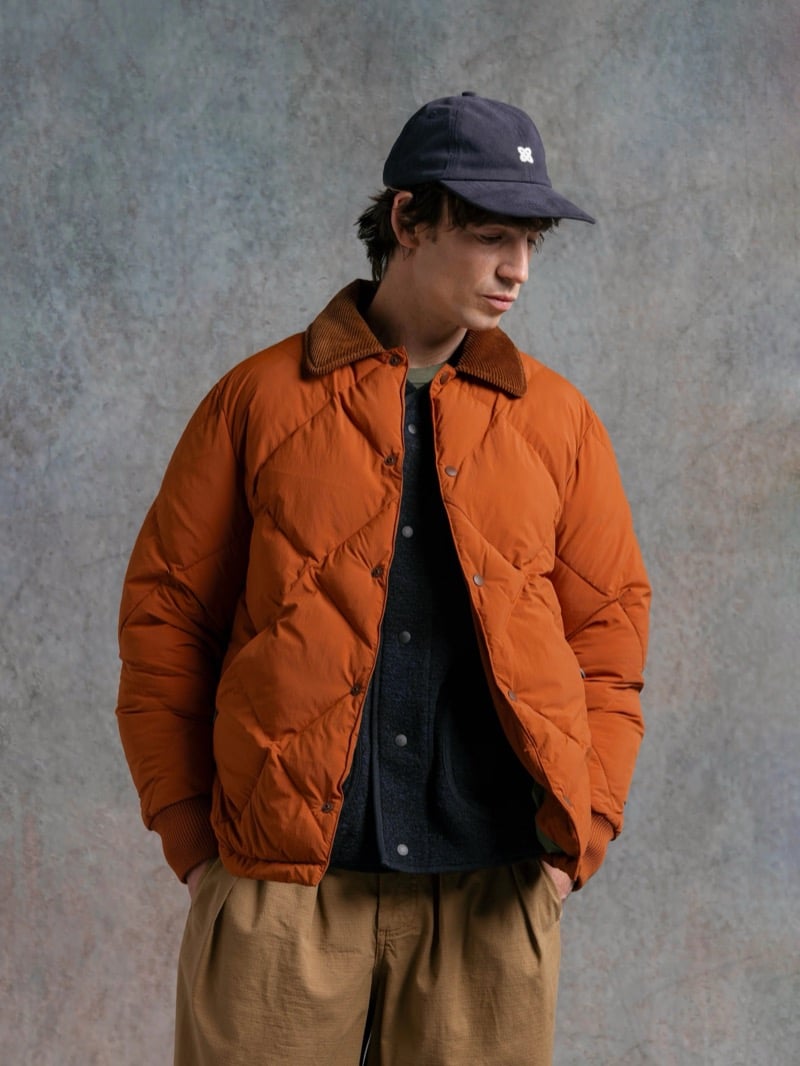 Man in a burnt orange quilted jacket over an olive sweater, wearing a cap, for Kestin Fall 2026