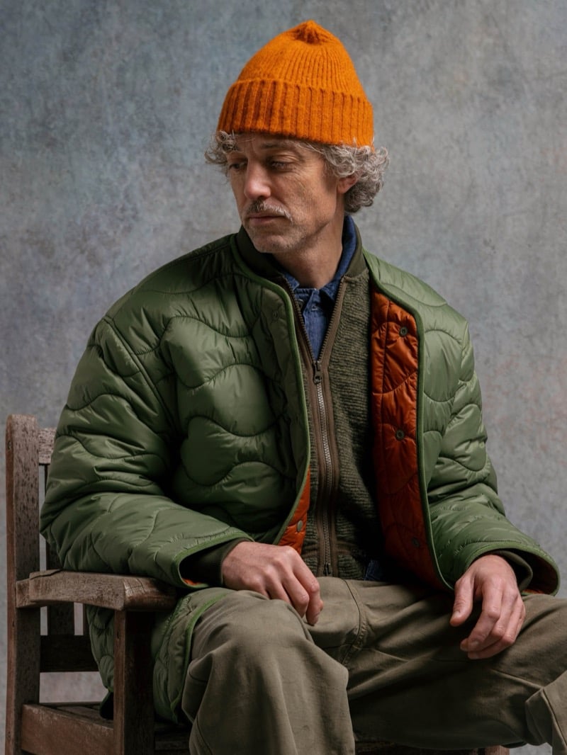 Man in an olive quilted jacket with an orange lining, wearing an orange beanie, for Kestin Fall 2026